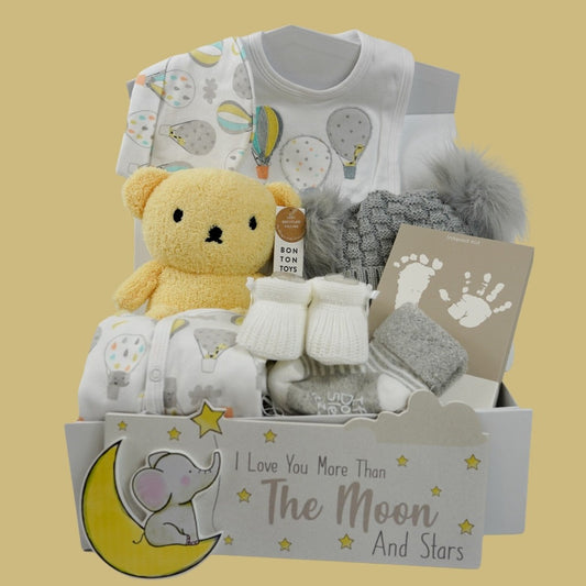 Baby hamper box with yellow Boris bear in terry fabric, white baby sleepsuit with colourful hot air balloons and matching hat, bib, hand and footprint ink pad, baby knit white booties, grey knit hat with double fluffy pom pom