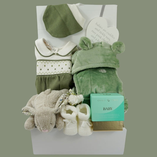 white hamper box with baby gifts including a green and cream smocked baby sleepsuit with matching hat, green baby dressing gown with cute ears, Soft bunny toy, Luxury Mumm and Baby candle, white baby booties, white nursery plaque 