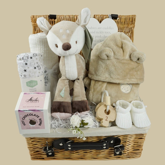 baby hamper in wicker basket with a fawn soft toy, white cotton cellular blanket, while soft large muslin with grey elephants and stars, soft fawn coloured dressing gown with cute teddy ears, white knit booties, set of wooden teething keys on a rope, hot chocolate bomb 
