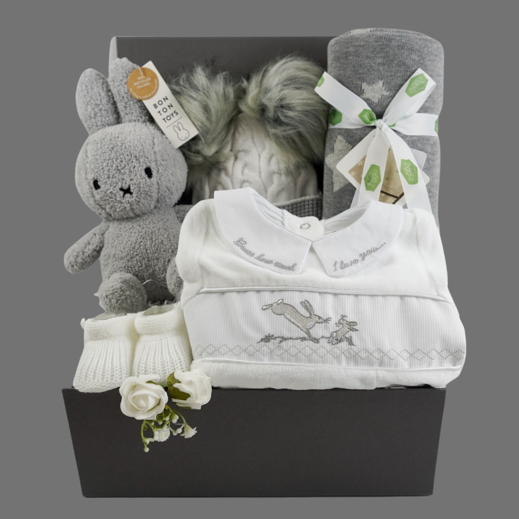 Grey hamper box with baby items includes white velour baby sleepsuit with Guess how much I love embroidered in grey on the collar and a smocked front with rabbits on the front, white knit baby booties, grey and white starred baby blanket, cream and grey knit baby hat with grey fluffy pom poms , grey terry Miffy rabbit
