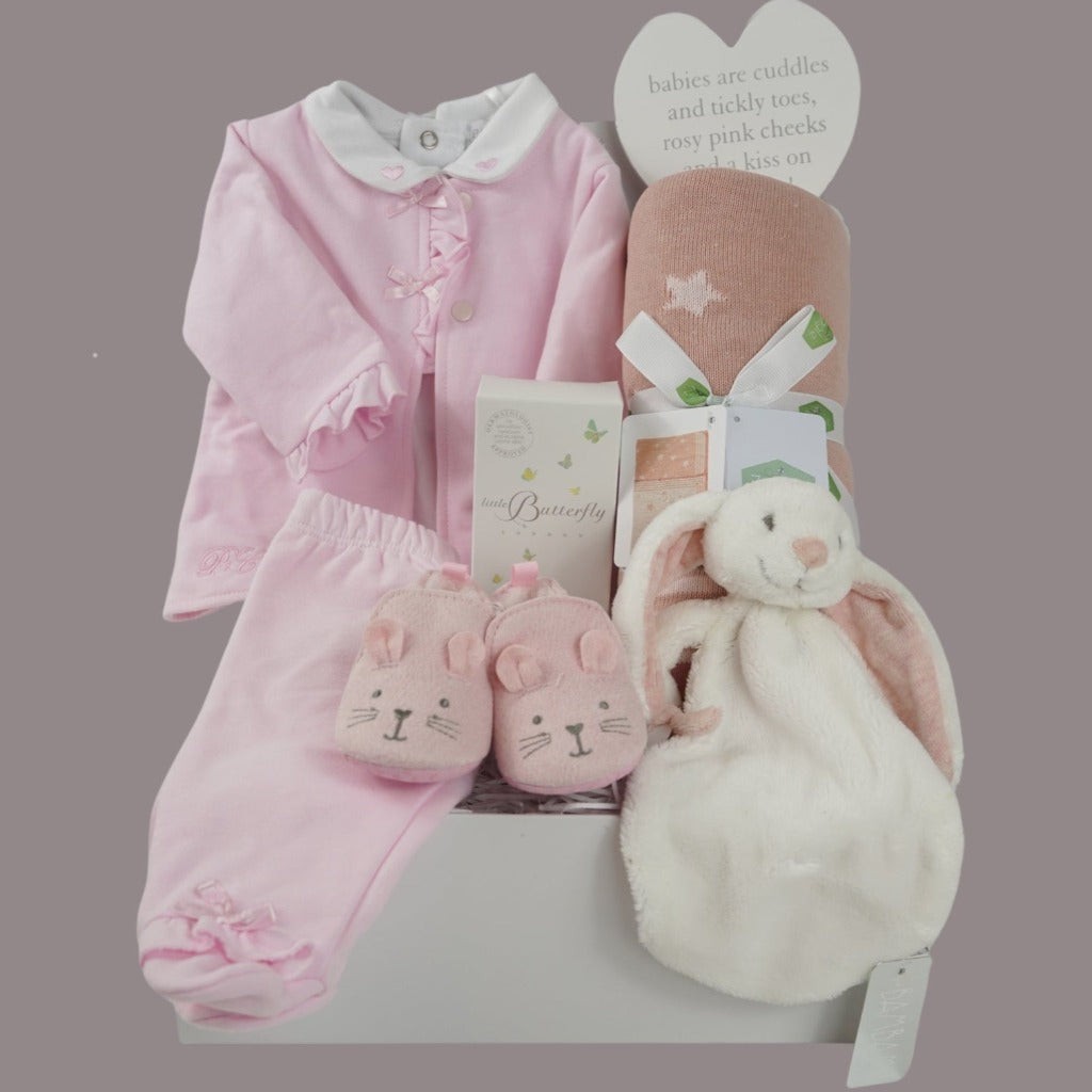 Baby hamper with pink and white baby outfit including a baby bodysuit in white with pink hearts embroidered on the collar, pink cardigan with frilled edges and bows and frilled cuffs and around the feet on the pink leggings, dusky pink star baby blanket, organic baby toiletries, white rabbit baby tuttle, pink baby shoes with a cute face and ears