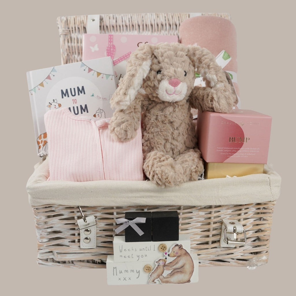 white wicker hamper basket with mum to be gifts includes box with toiletries for mum and baby, mum to mum advice book, pale pink ribbed baby sleepsuit with zip fastening, soft carmel coloured bunny rabbit soft toy, dusky pink heavy weight baby blanket with stars, bump pregnancy candle