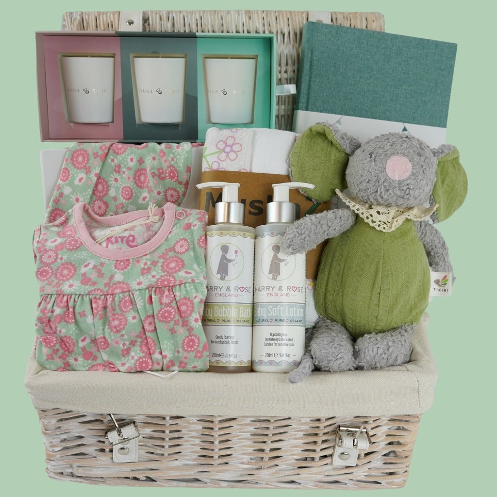 White hamper basket with baby girl organic gifts, pale green with pink flowers baby organic sleepsuit , matching organic hat, organic baby wash and body lotion, organic muslins in white with pink flowers, organic mouse soft toy, The story of us baby journal,  motherhood 3 mini candles