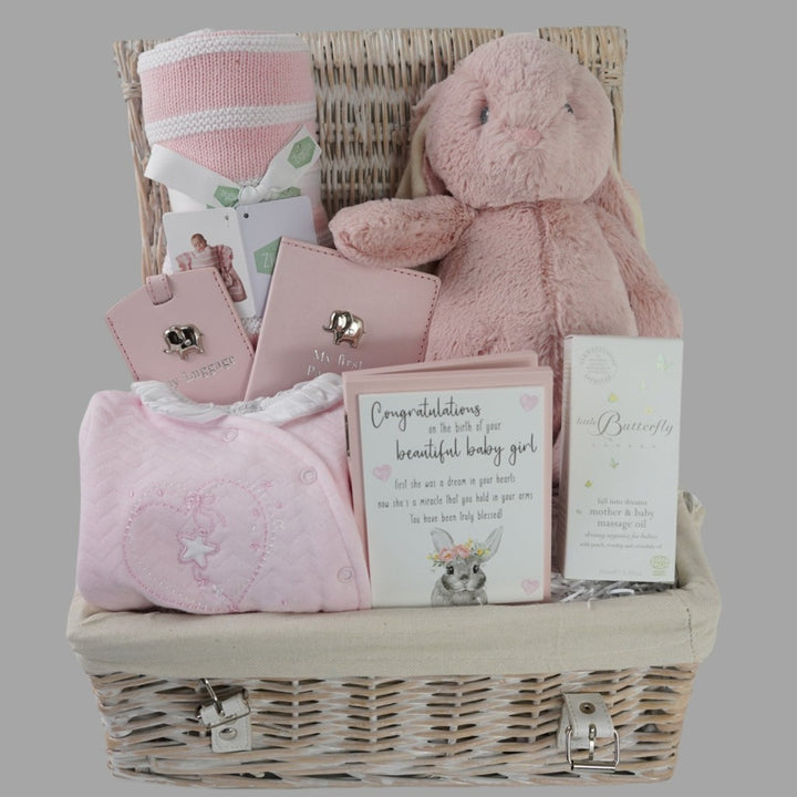 White hamper basket with baby girl gifts including pink baby quilted outfit with white frilled collar, baby pink passport holder and luggage label with silver elephant, pink and white heavy knit baby blanket, pink soft cuddly eco friendly bunny, little butterfly london organic baby and mum massage oil in a box, wooden forever congratulation card