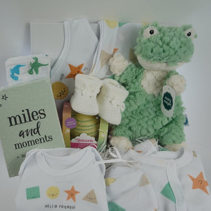 white hamper box with baby gifts including a white cotton layette set with shapes designs on the sleepsuit and vest, baby milestone cards in a green box, white hot chocolate bomb , white muslin with green and blue dinosaurs, white knit baby booties , green frog lovey