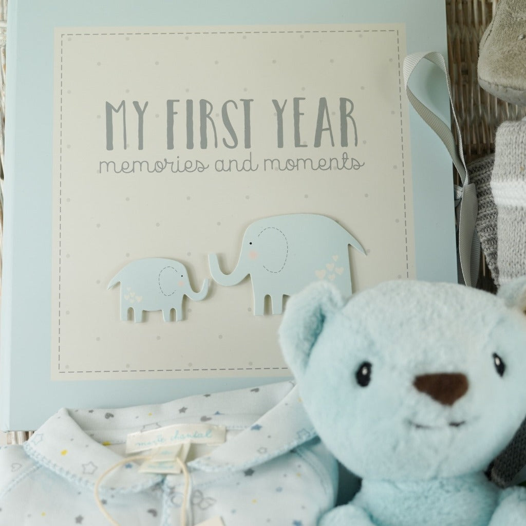 Luxury baby hamper in a wicker basket, Blue steiff my first teddy, Star and Crown baby sleepsuit in blue, My First year blue baby book, grey baby suede look slippers with cute face, grey and white heave baby blanket, forever baby sentiment card in blue with elephant design