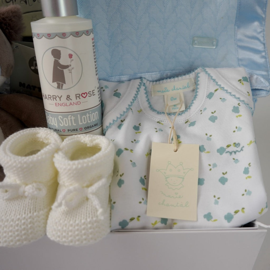 baby boy hamper box, newborn baby bodysuit with long sleeves and matching baby hat, blue baby wrap with satin ribbon edging, musical racoon soft toy, congratulations chocolates, white knit booties and baby body lotion