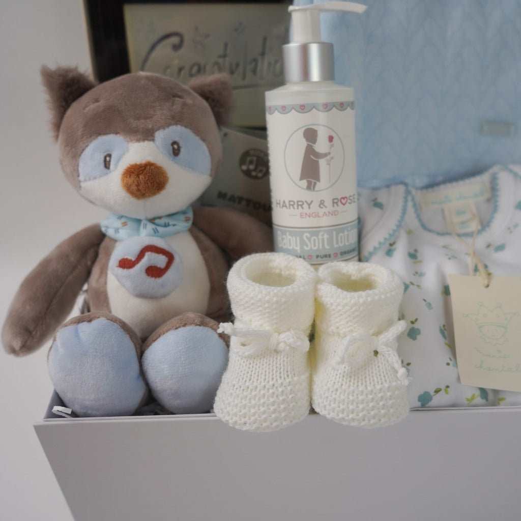 baby boy hamper box, newborn baby bodysuit with long sleeves and matching baby hat, blue baby wrap with satin ribbon edging, musical racoon soft toy, congratulations chocolates, white knit booties and baby body lotion