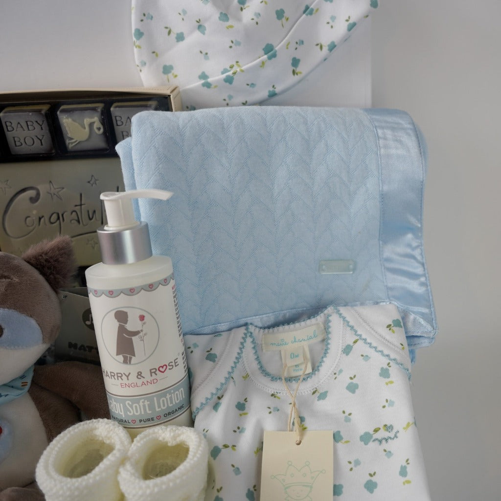 baby boy hamper box, newborn baby bodysuit with long sleeves and matching baby hat, blue baby wrap with satin ribbon edging, musical racoon soft toy, congratulations chocolates, white knit booties and baby body lotion