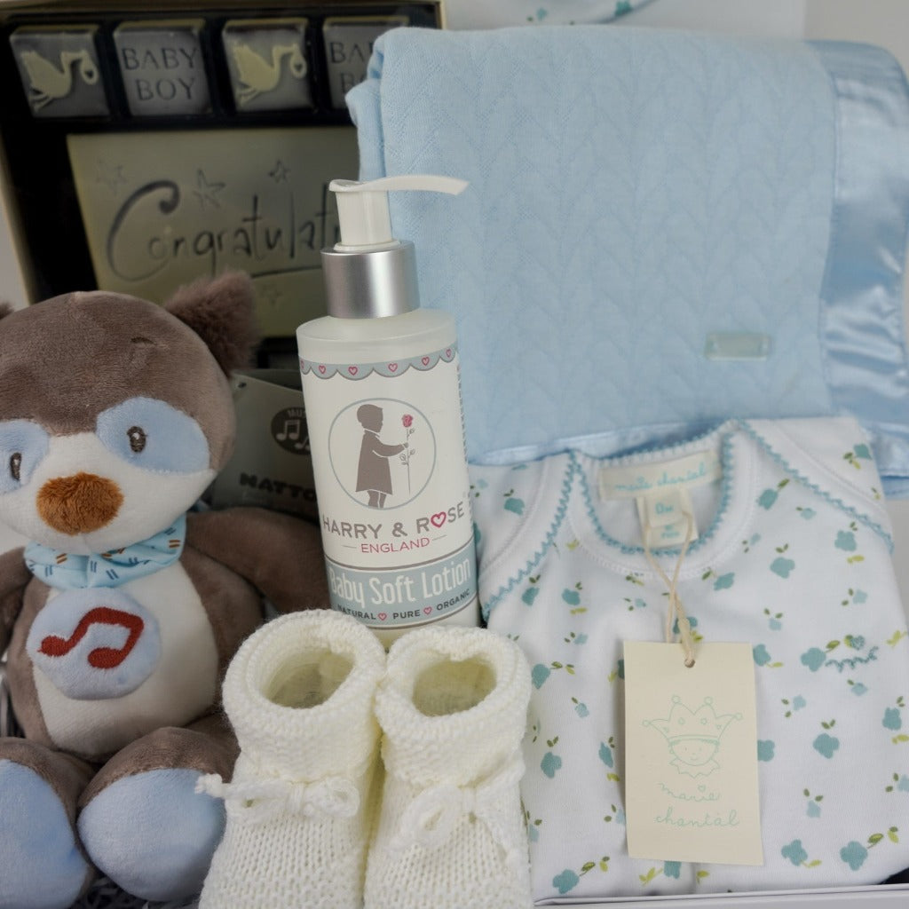 baby boy hamper box, newborn baby bodysuit with long sleeves and matching baby hat, blue baby wrap with satin ribbon edging, musical racoon soft toy, congratulations chocolates, white knit booties and baby body lotion