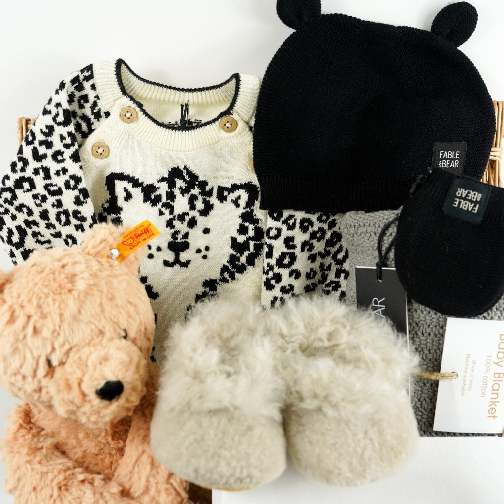 baby hamper basket with honey coloured steiff teddy, baby alpaca slippers in champagne, baby knitted jumper in organic cream cotton with black leopard design, black baby cotton knitted leggings, black cotton knit baby hat with ears, baby photo album in white linen, baby stacking ring toy with rabbit head, grey cotton baby cellular blanket