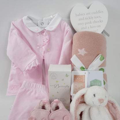 Baby hamper with pink and white baby outfit including a baby bodysuit in white with pink hearts embroidered on the collar, pink cardigan with frilled edges and bows and frilled cuffs and around the feet on the pink leggings, dusky pink star baby blanket, organic baby toiletries, white rabbit baby tuttle, pink baby shoes with a cute face and ears