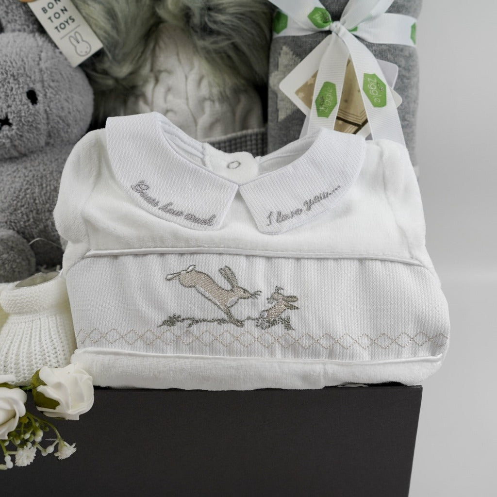 Grey hamper box with baby items includes white velour baby sleepsuit with Guess how much I love embroidered in grey on the collar and a smocked front with rabbits on the front, white knit baby booties, grey and white starred baby blanket, cream and grey knit baby hat with grey fluffy pom poms , grey terry Miffy rabbit