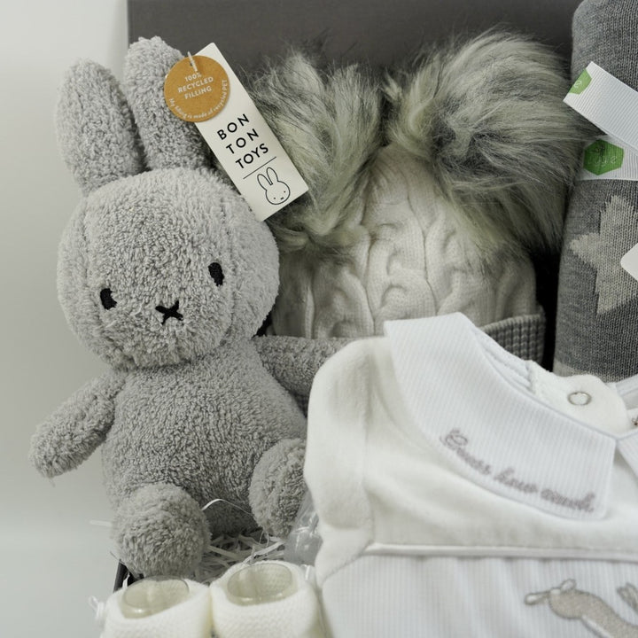 Grey hamper box with baby items includes white velour baby sleepsuit with Guess how much I love embroidered in grey on the collar and a smocked front with rabbits on the front, white knit baby booties, grey and white starred baby blanket, cream and grey knit baby hat with grey fluffy pom poms , grey terry Miffy rabbit