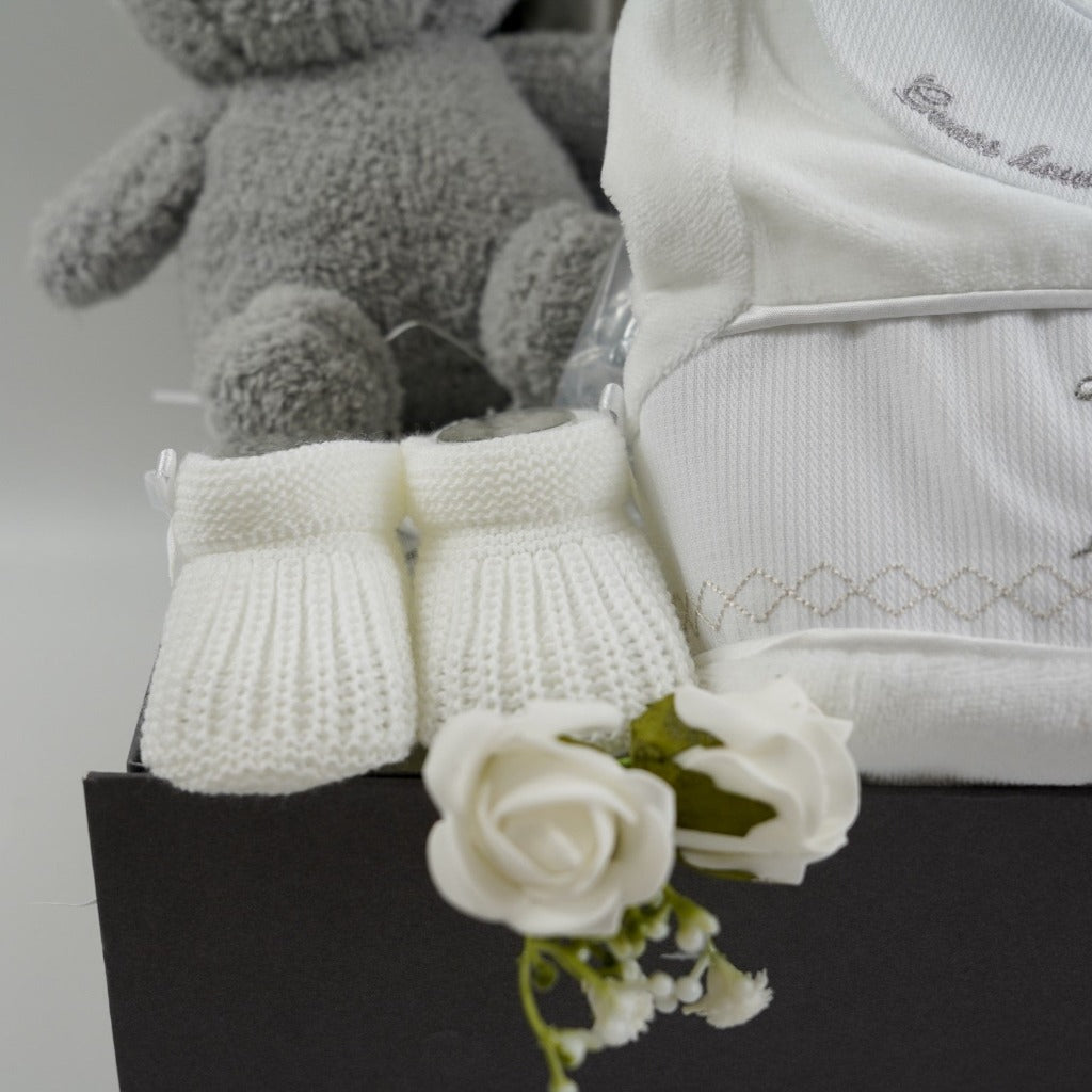 Grey hamper box with baby items includes white velour baby sleepsuit with Guess how much I love embroidered in grey on the collar and a smocked front with rabbits on the front, white knit baby booties, grey and white starred baby blanket, cream and grey knit baby hat with grey fluffy pom poms , grey terry Miffy rabbit