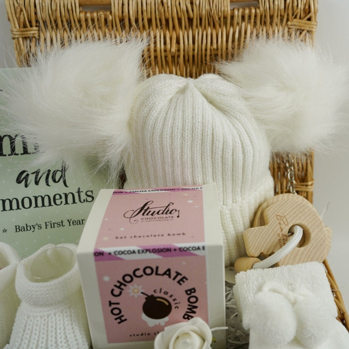 Small hamper basket with baby milestone cards in a box, white knit hat with 2 fluffy pom poms, baby booties in white, white baby socks with a cute message on the soles, wooden teething keys , Hot chocolate bomb