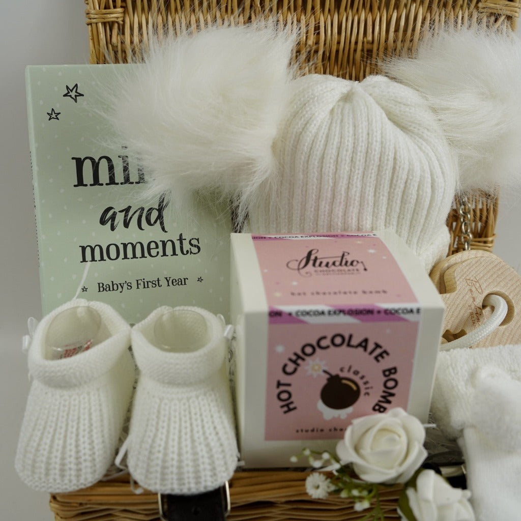 Small hamper basket with baby milestone cards in a box, white knit hat with 2 fluffy pom poms, baby booties in white, white baby socks with a cute message on the soles, wooden teething keys , Hot chocolate bomb