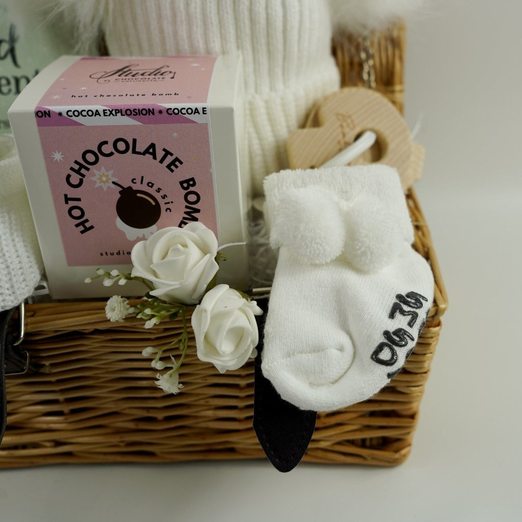Small hamper basket with baby milestone cards in a box, white knit hat with 2 fluffy pom poms, baby booties in white, white baby socks with a cute message on the soles, wooden teething keys , Hot chocolate bomb