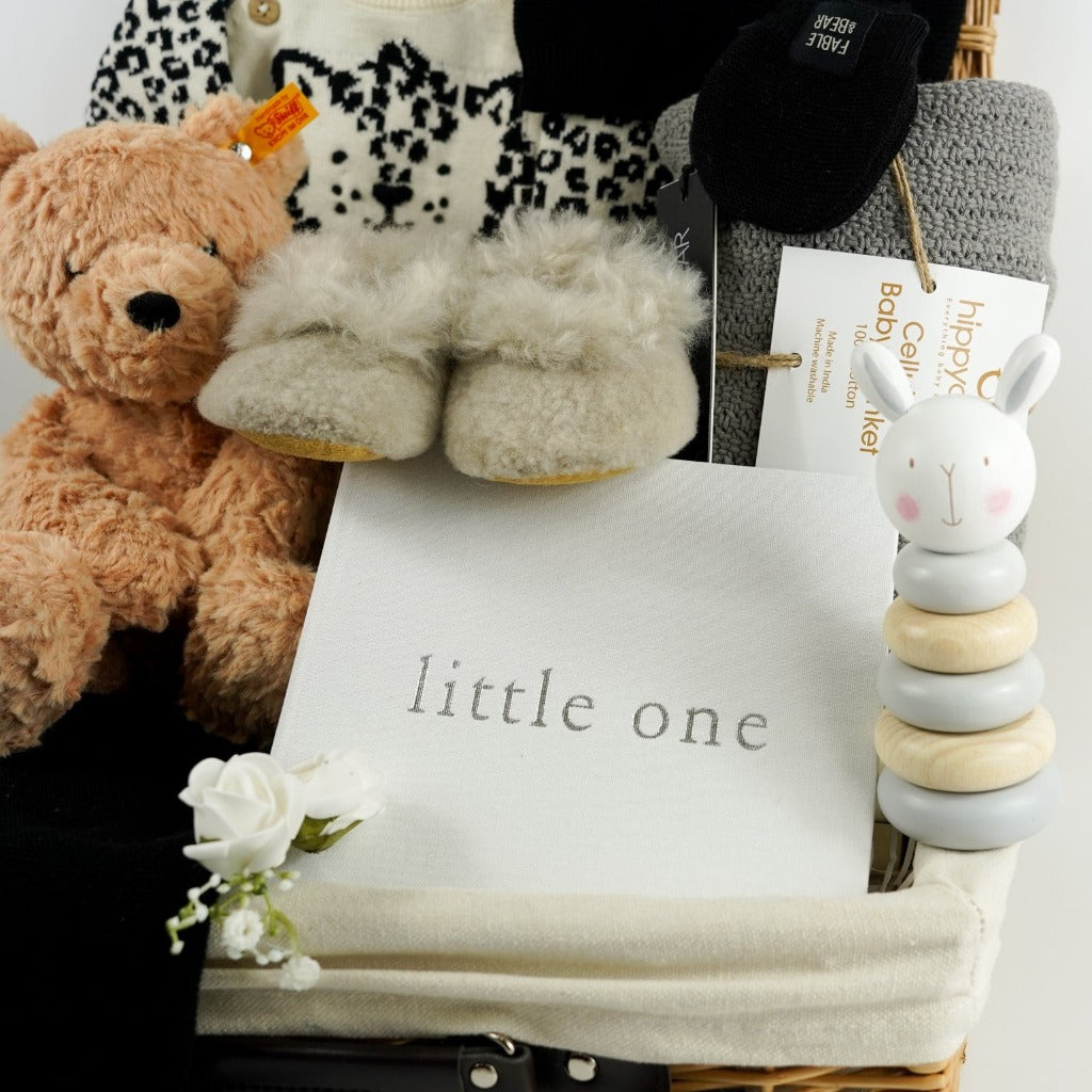 baby hamper basket with honey coloured steiff teddy, baby alpaca slippers in champagne, baby knitted jumper in organic cream cotton with black leopard design, black baby cotton knitted leggings, black cotton knit baby hat with ears, baby photo album in white linen, baby stacking ring toy with rabbit head, grey cotton baby cellular blanket