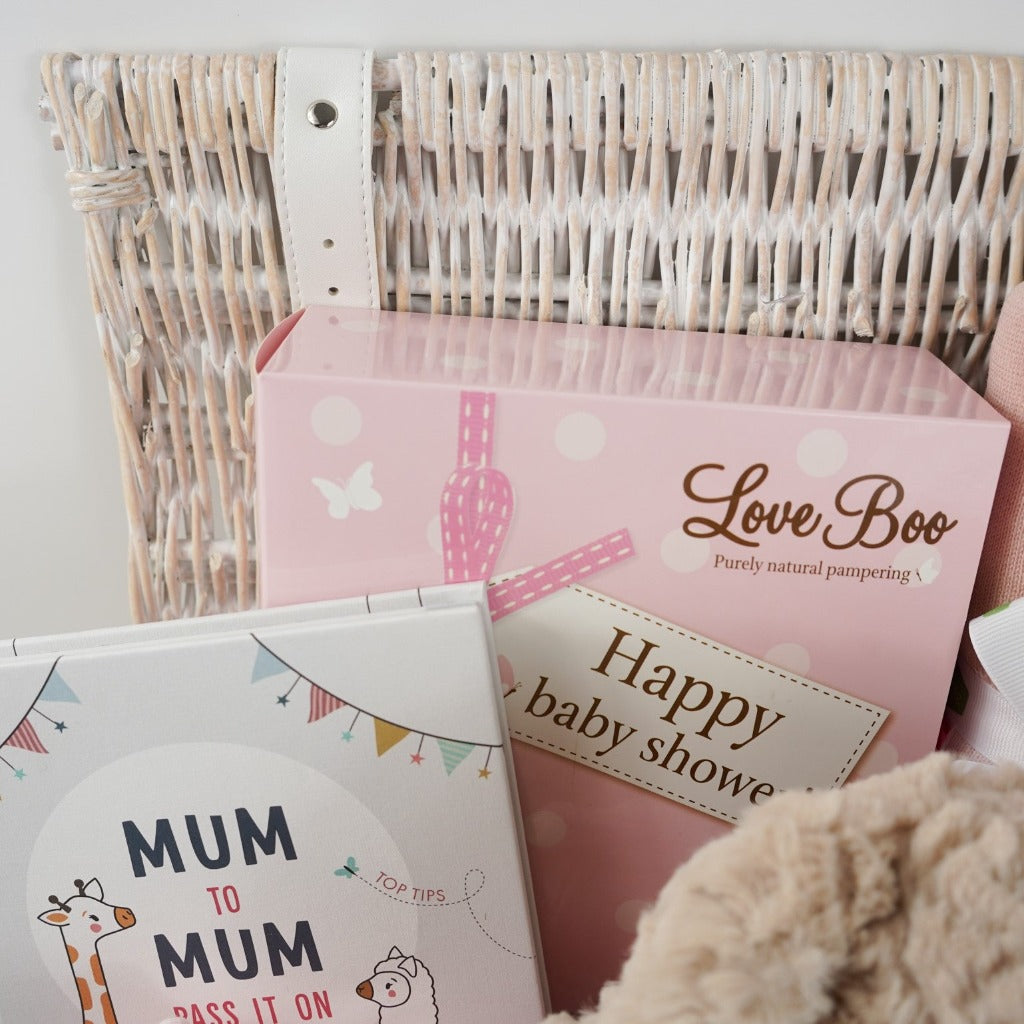 white wicker hamper basket with mum to be gifts includes box with toiletries for mum and baby, mum to mum advice book, pale pink ribbed baby sleepsuit with zip fastening, soft carmel coloured bunny rabbit soft toy, dusky pink heavy weight baby blanket with stars, bump pregnancy candle