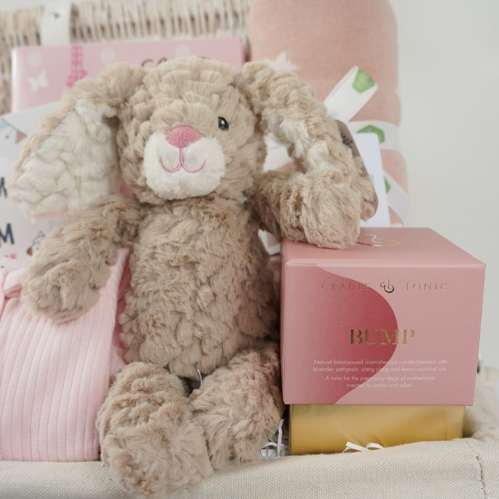 white wicker hamper basket with mum to be gifts includes box with toiletries for mum and baby, mum to mum advice book, pale pink ribbed baby sleepsuit with zip fastening, soft carmel coloured bunny rabbit soft toy, dusky pink heavy weight baby blanket with stars, bump pregnancy candle