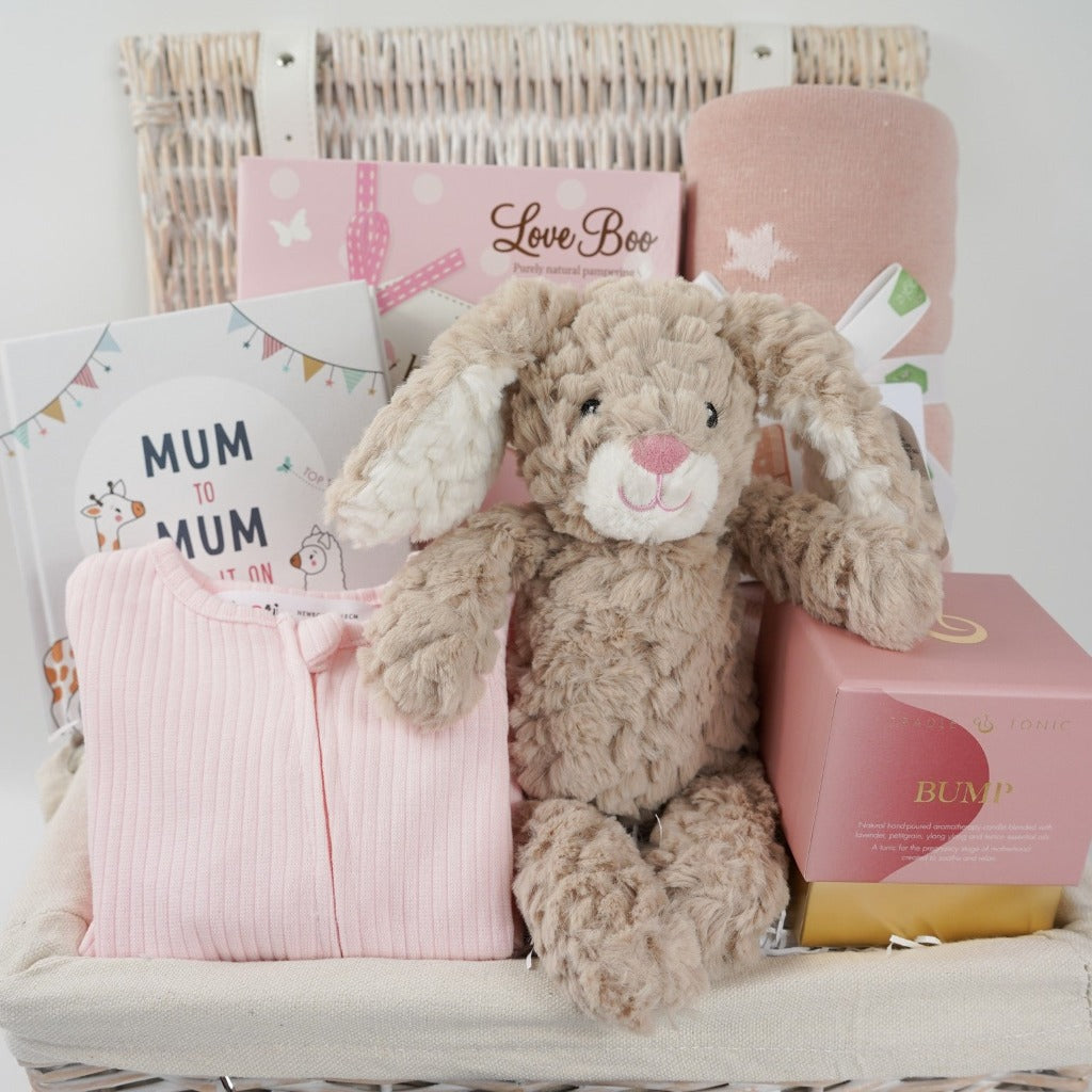 white wicker hamper basket with mum to be gifts includes box with toiletries for mum and baby, mum to mum advice book, pale pink ribbed baby sleepsuit with zip fastening, soft carmel coloured bunny rabbit soft toy, dusky pink heavy weight baby blanket with stars, bump pregnancy candle