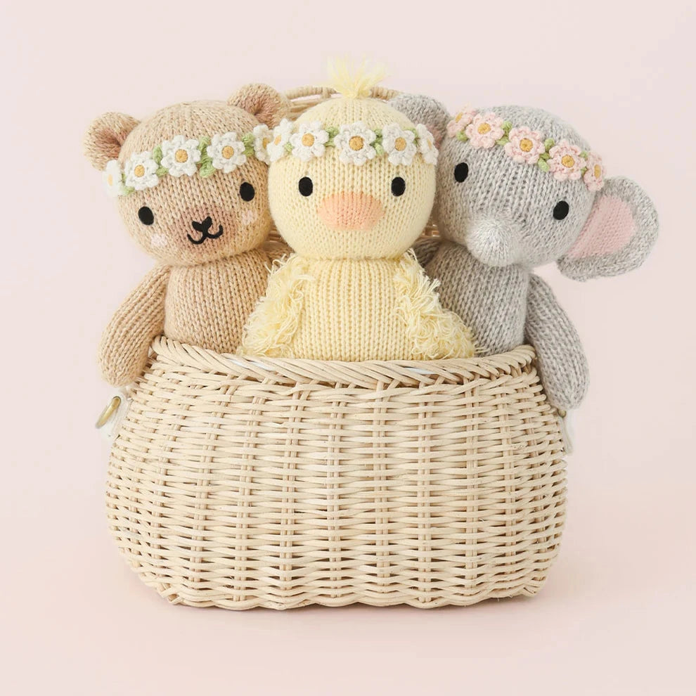 Three knitted toys, including a bear, a rabbit, and a chick, sitting in a woven basket on a light pink background.