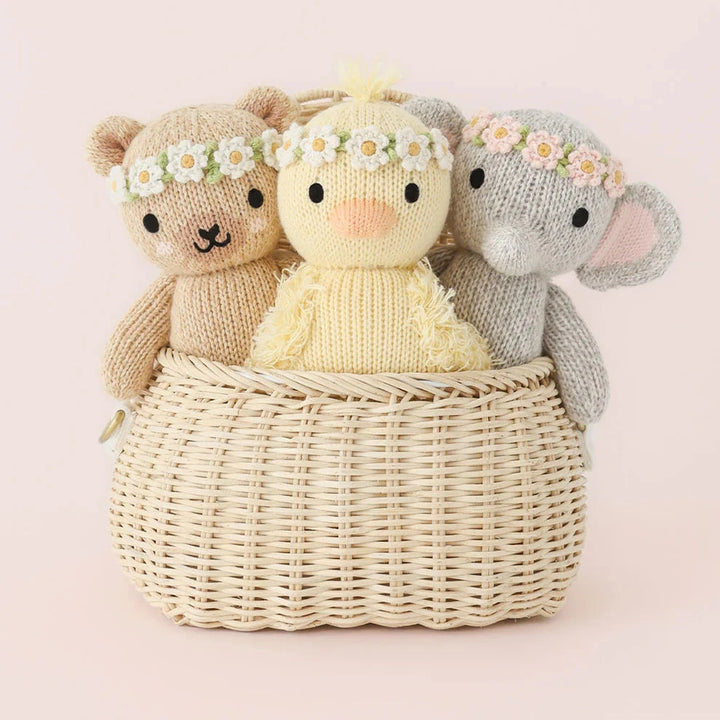 Three knitted toys, including a bear, a rabbit, and a chick, sitting in a woven basket on a light pink background.