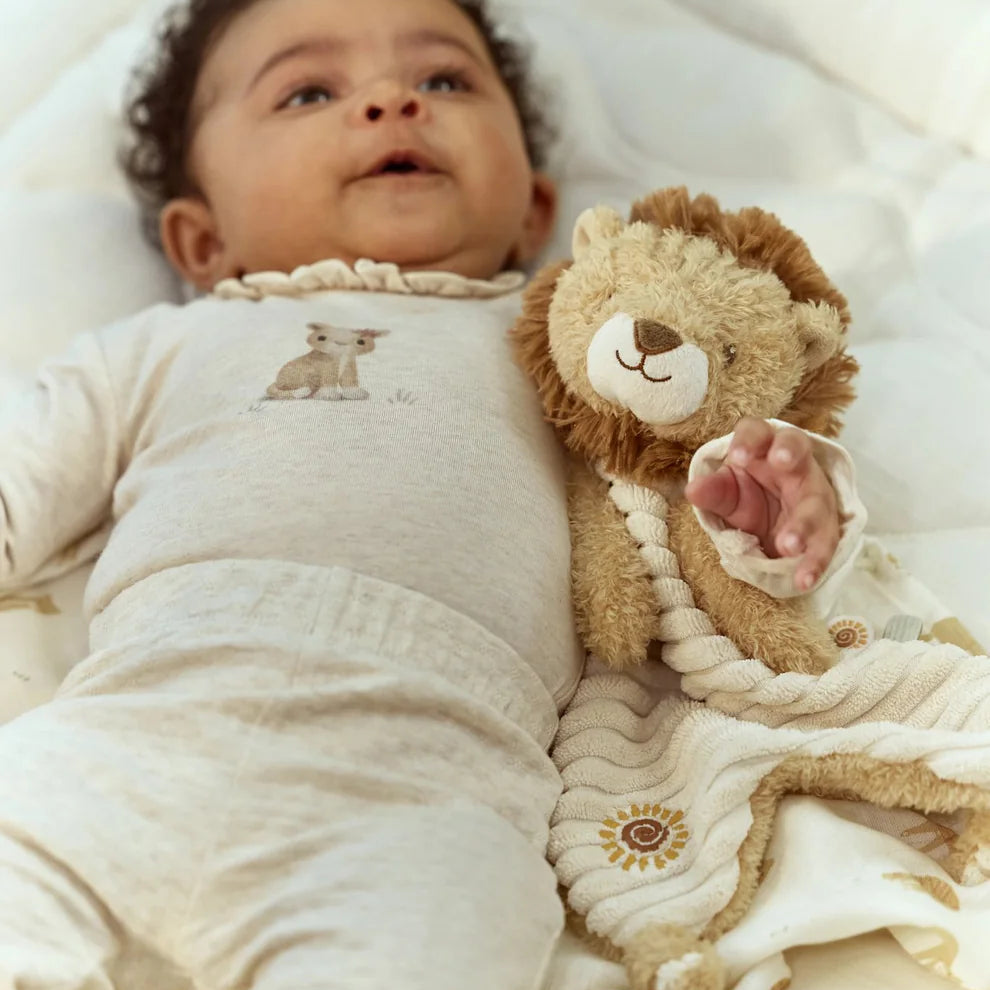 A baby lying in a bassinet wearing a cream-colored footed sleeper with a small animal graphic on the chest, next to a plush teddy bear and a soft blanket.