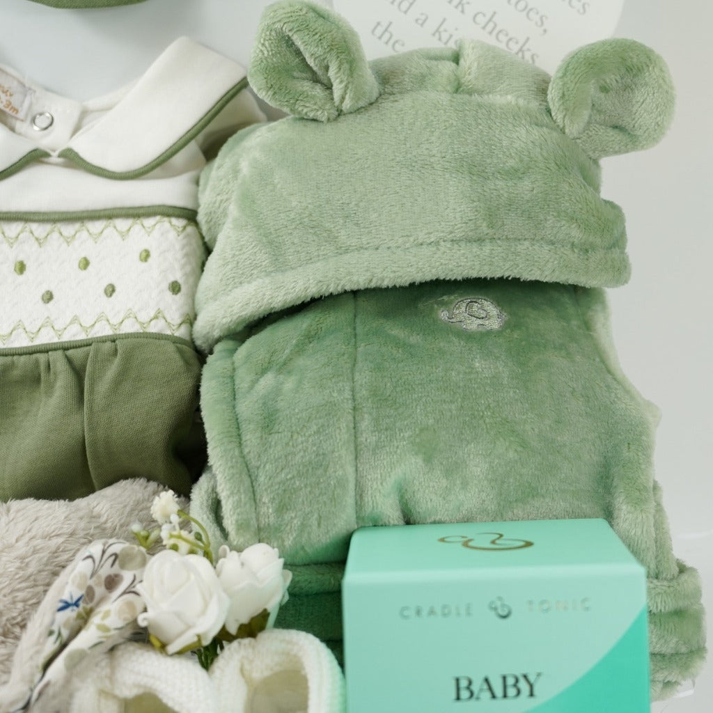 white hamper box with baby gifts including a green and cream smocked baby sleepsuit with matching hat, green baby dressing gown with cute ears, Soft bunny toy, Luxury Mumm and Baby candle, white baby booties, white nursery plaque