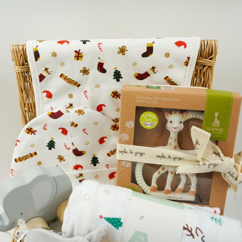 luxury cream baby christmas bib and hat set with Christmas trees, crackers, presents and stockings design, Sophie la Girafe teether, white muslin with reindeer and christmas trees, wooden push along elephant, Steiff crincle elephant rattle all in a small hamper basket