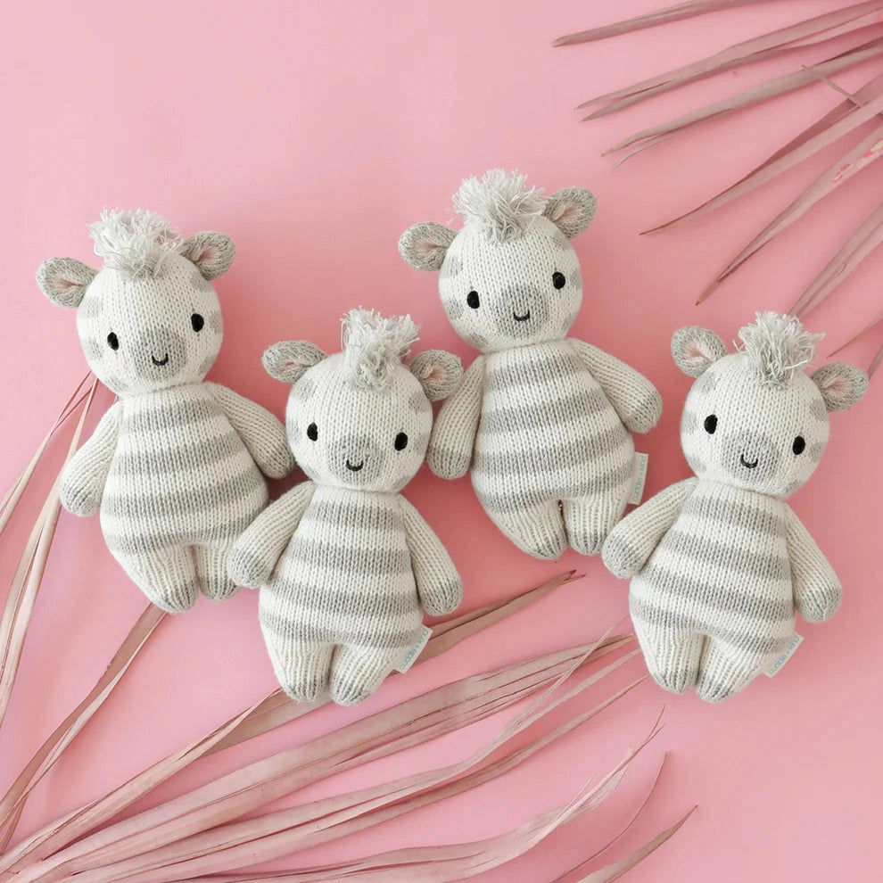 Four knitted baby toys shaped like zebras on a pink background with dried leaves.