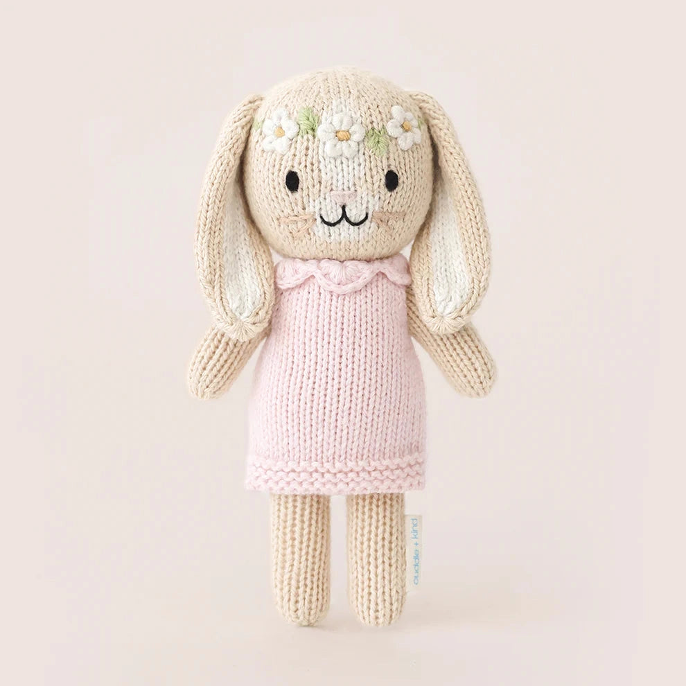 A knitted bunny plush toy with long floppy ears, a cream-colored body, a light pink dress, and small white daisy flowers on its forehead, set against a solid pale pink background.
