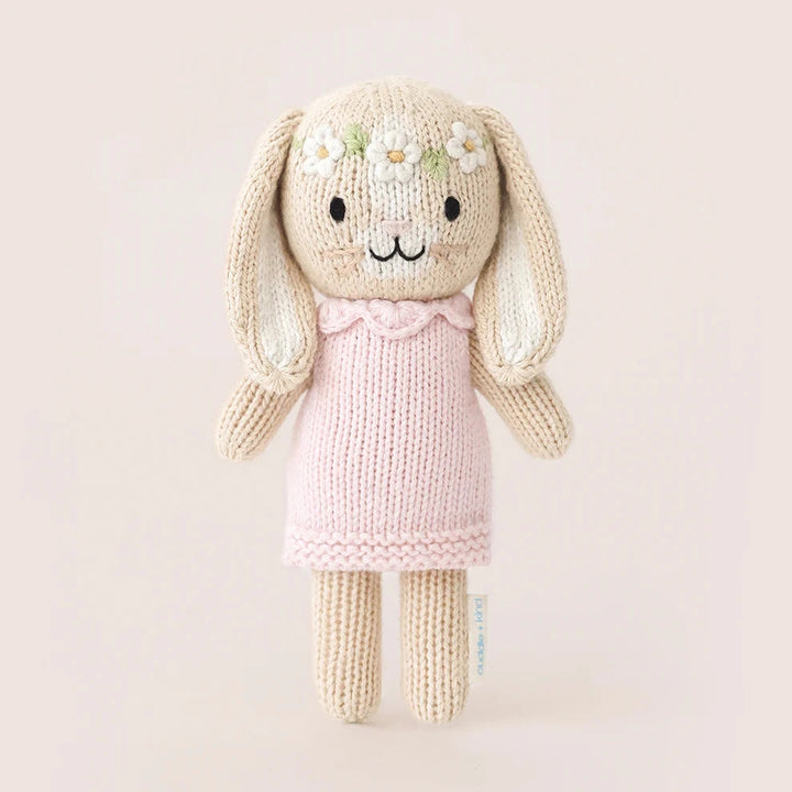 A knitted bunny plush toy with long floppy ears, a cream-colored body, a light pink dress, and small white daisy flowers on its forehead, set against a solid pale pink background.