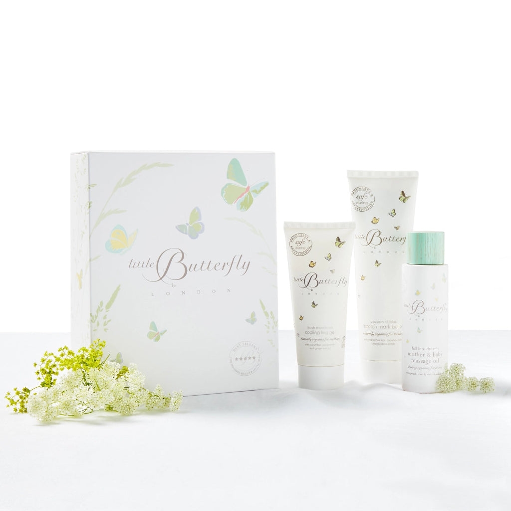 A pregnancy skincare gift set from Little Butterfly London featuring a white 'little Butterfly' box and three matching white tubes and bottles with delicate butterfly illustrations, green accents, and small white flowers placed around them 