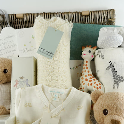 Luxury baby gifts in a hamper basket, Luxury New Mum candle , Angel Wings 2 piece baby clothing set in cream with gold angel wing design, baby hairbrush, baby cashmere shawl, sophie la giraffe, organic steiff teddy and comforter, baby jourmal , baby slippers and baby swaddle in white with grey safari animals
