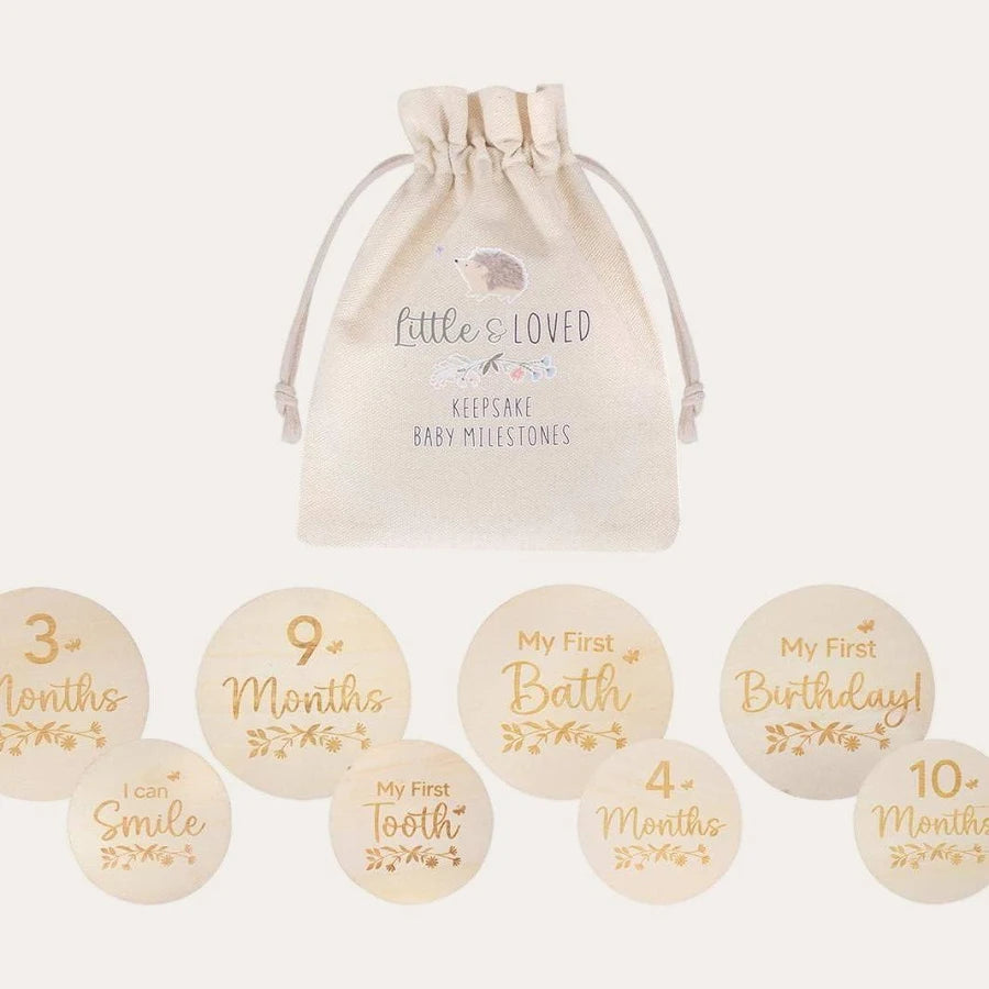 A set of round baby milestone stickers with gold lettering on a cream fabric base, displayed below a matching cream drawstring bag printed with 'Little One LOVED Keepsake Baby Milestones'.