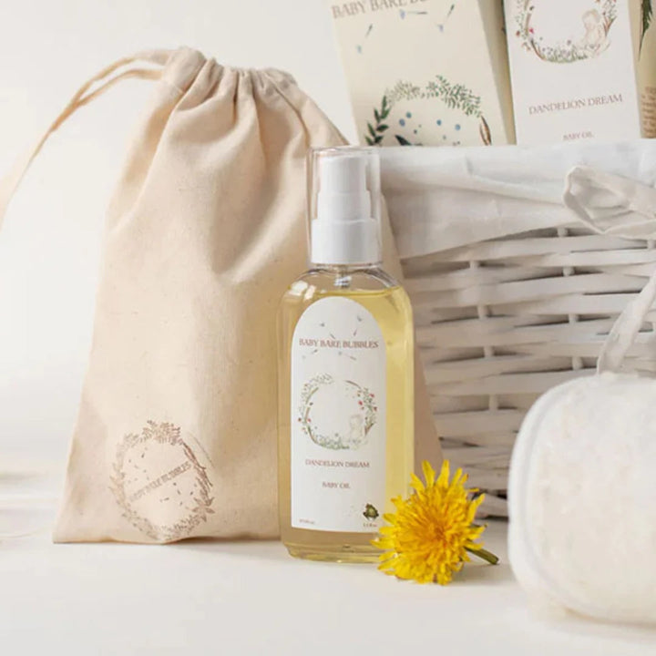 Baby Baze Blender Ginger Cream body oil in a clear spray bottle, styled with a beige drawstring pouch, a white wicker basket, a white bath sponge, and a small yellow flower on a white surface.