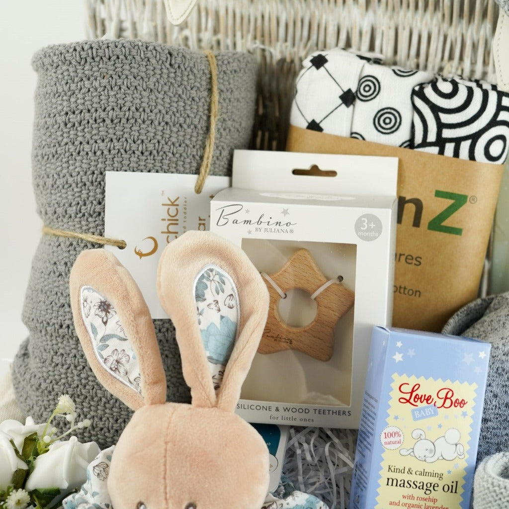 White wash wicker hamper with baby items, Doudou et Compagnie rabbit comforter in blue, grey bamboo cellular blanket, sensory black and white muslins, milestone cards, Aden and Anais baby grey gown with knot hat, wooden star teether, baby knit grey bootees, baby massage oil
