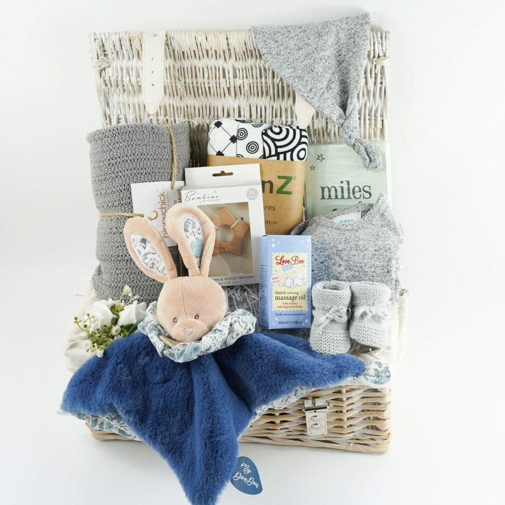 White wash wicker hamper with baby items, Doudou et Compagnie rabbit comforter in blue, grey bamboo cellular blanket, sensory black and white muslins, milestone cards, Aden and  Anais baby grey gown with knot hat, wooden star teether, baby knit grey bootees, baby massage oil