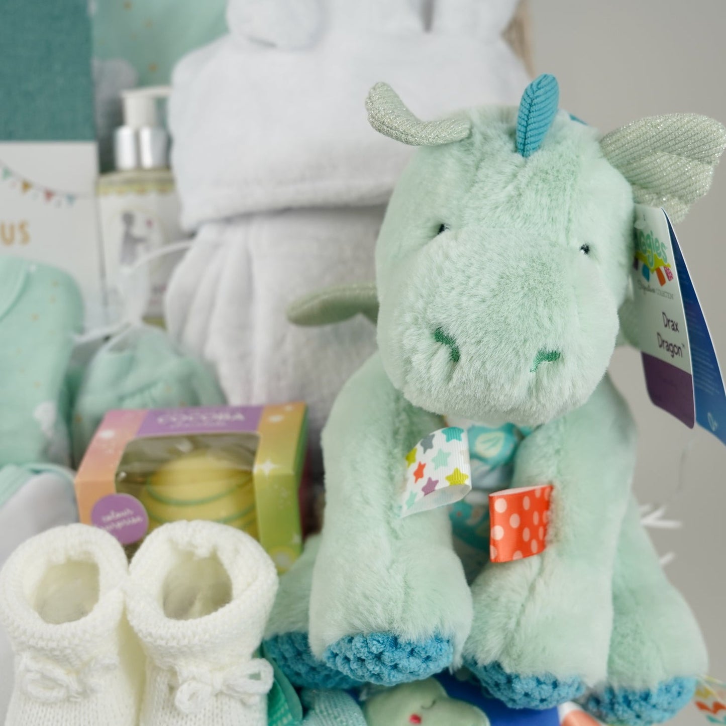 baby hamper basket, baby journal, baby layette outfit in pale mint green with elephants , white baby dressing gown with cute ears, white hot chocolate bomb, baby organic bubble bath, Dragon taggie in green and matching taggie book , white knit booties