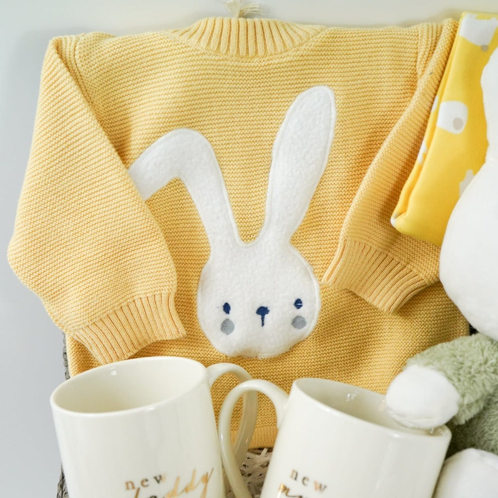 baby hamper in yellow organic knit with a white bunny face, White Miffy soft toy with green fluffy outfit and matching Miffy comforter , Cream bone china new mummy and new daddy mugs, yellow hat with cute ears and bunny , white booties