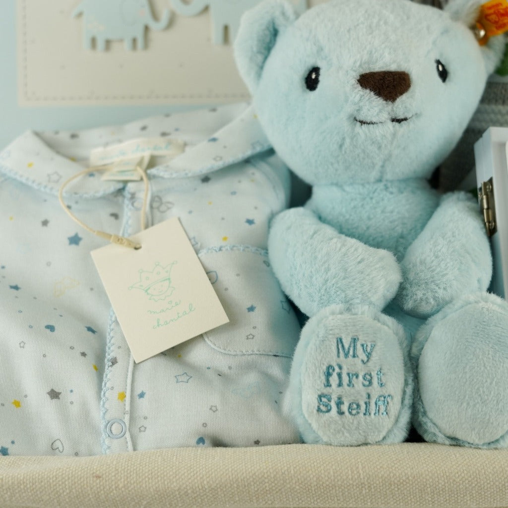Luxury baby hamper in a wicker basket, Blue steiff my first teddy, Star and Crown baby sleepsuit in blue, My First year blue baby book, grey baby suede look slippers with cute face, grey and white heave baby blanket, forever baby sentiment card in blue with elephant design