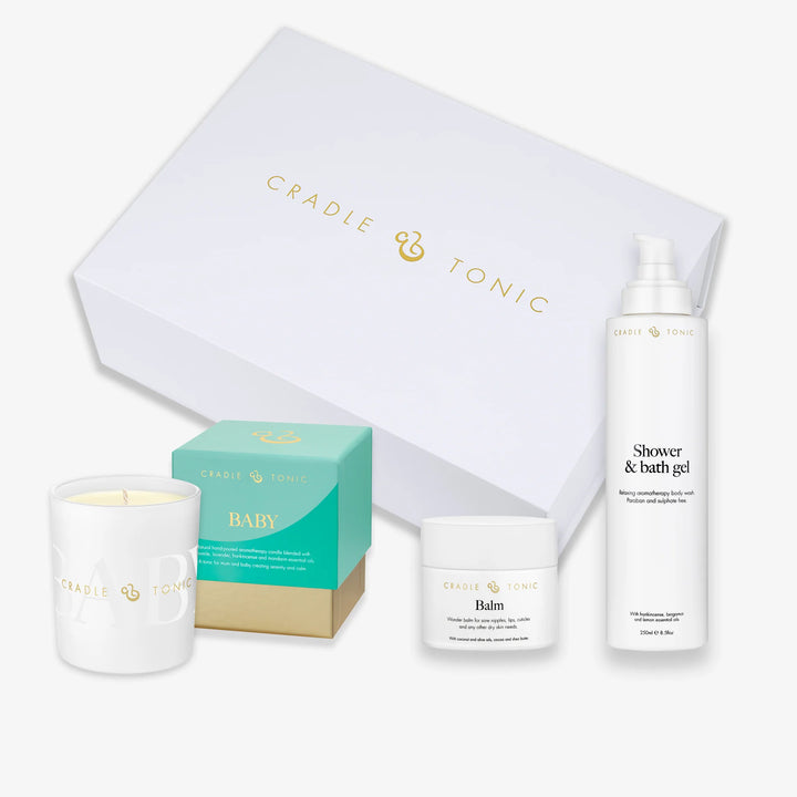 Cradle & Tonic baby gift set featuring a white scented candle, shower gel, balm, and a teal and gold BABY box, arranged in front of a large white gift box on a light gray background.