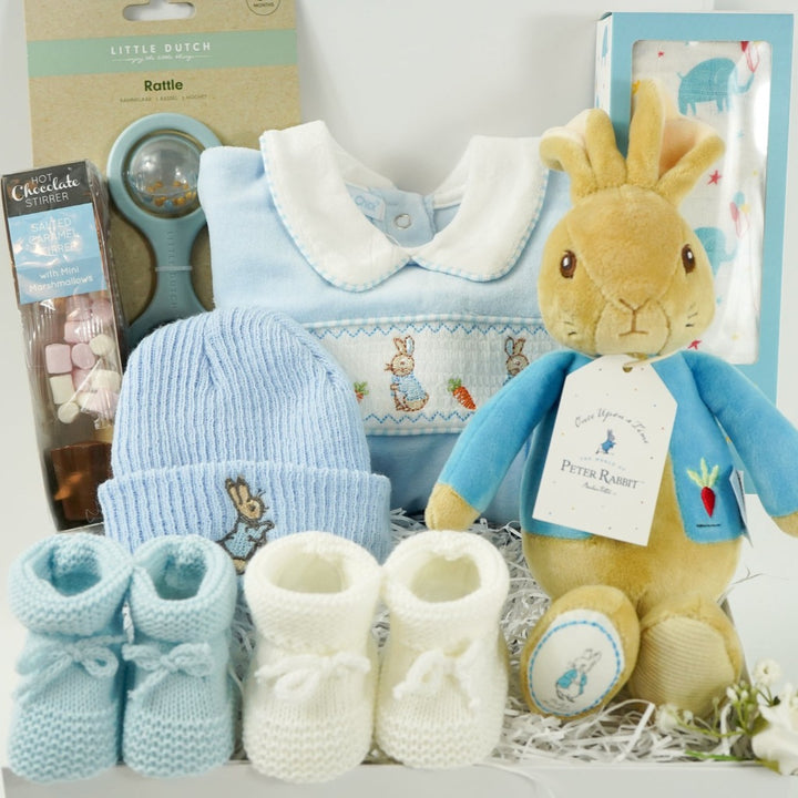 white baby hamper box with Beatrix potter Peter rabbit soft toy, blue velour baby sleepsuit with smocked yolk with bunny's , blue beanie hat with bunny, Little Dutch baby rattle, white muslin swaddle with blue elephants, salted caramel chocolate spoon with marshmallow