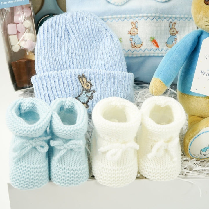 white baby hamper box with Beatrix potter Peter rabbit soft toy, blue velour baby sleepsuit with smocked yolk with bunny's , blue beanie hat with bunny, Little Dutch baby rattle, white muslin swaddle with blue elephants, salted caramel chocolate spoon with marshmallow