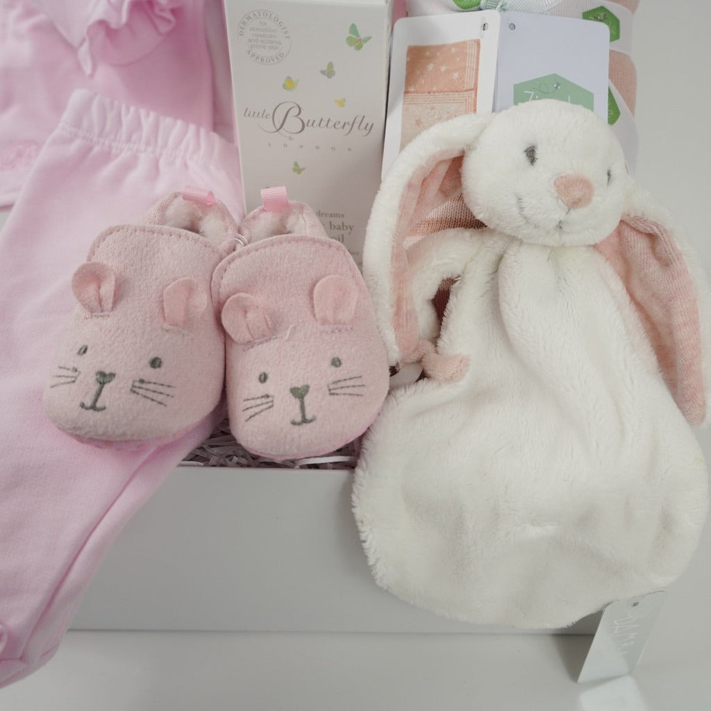 Baby hamper with pink and white baby outfit including a baby bodysuit in white with pink hearts embroidered on the collar, pink cardigan with frilled edges and bows and frilled cuffs and around the feet on the pink leggings, dusky pink star baby blanket, organic baby toiletries, white rabbit baby tuttle, pink baby shoes with a cute face and ears