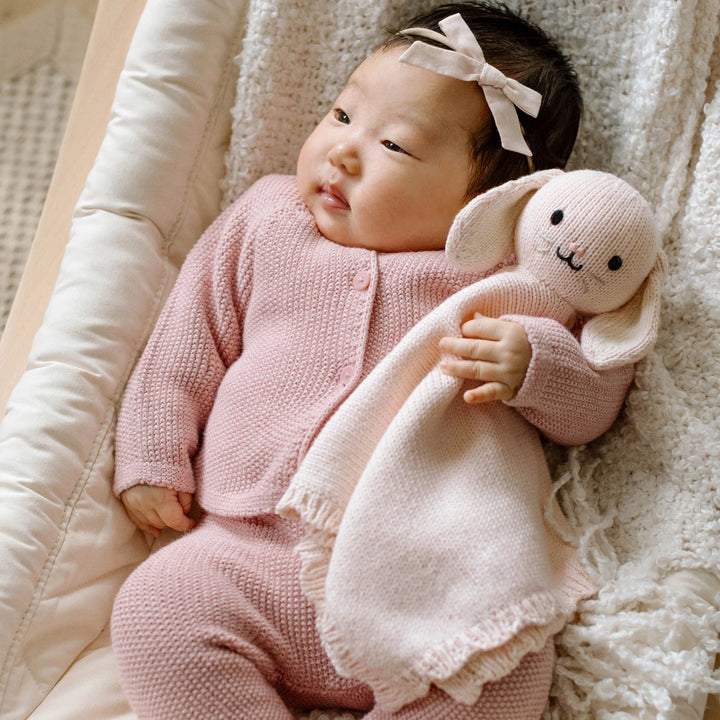 Baby in pink outfit holding a plush toy on a soft surface