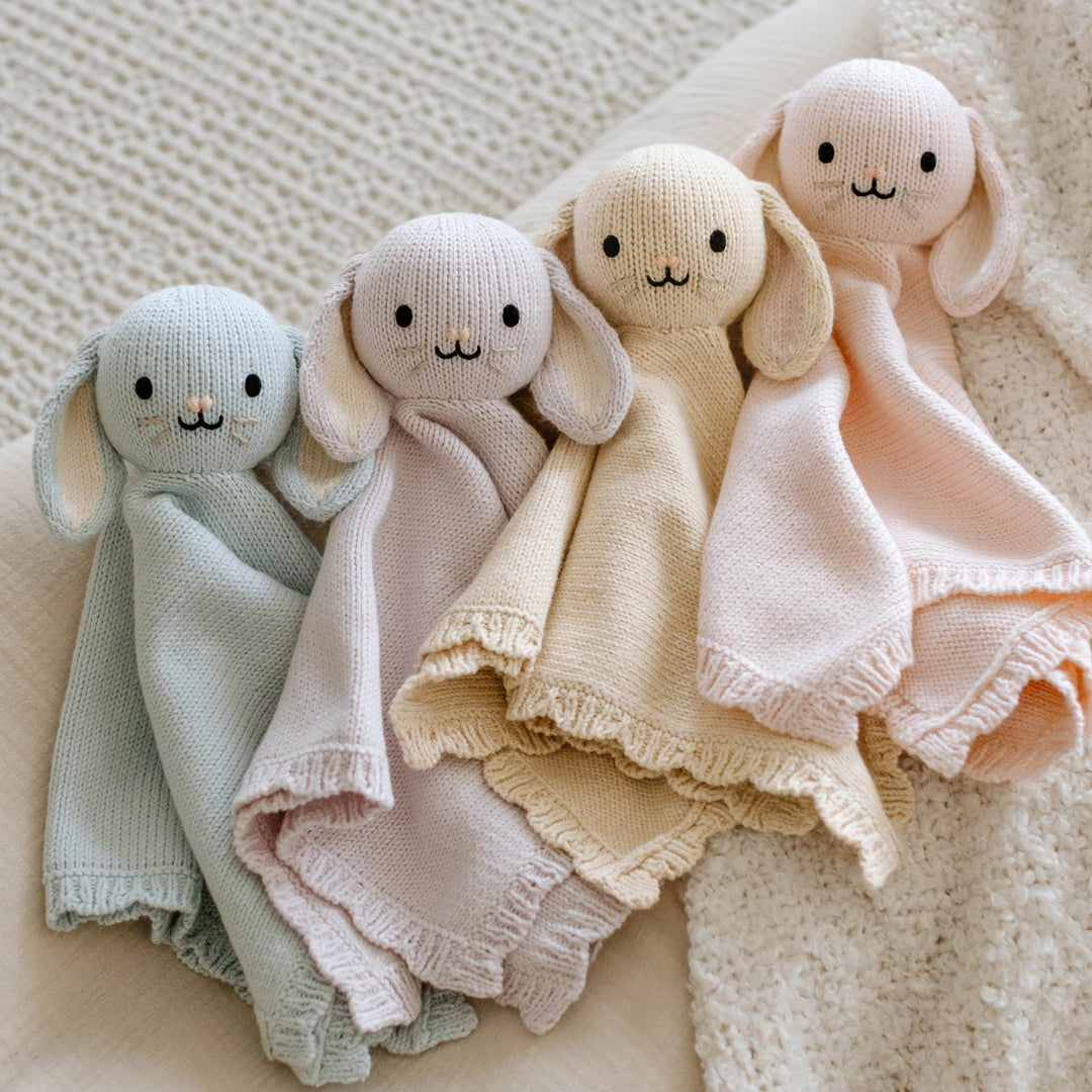 Four knitted baby comfort blankets in different colors on a textured surface
