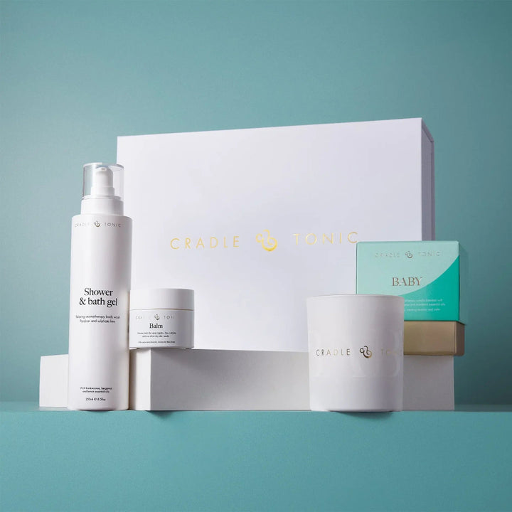 Cradie Tonic baby skincare gift set featuring a shower & bath gel bottle, a balm jar, a candle, and boxes on a teal background.